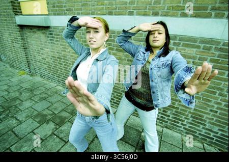Danielle Toele, IJmuiden, IJmuiden, Paesi Bassi, 20-05-2003 Whizgle News, Dutch Desk, Paesi Bassi, 1950-2000 due giovani donne colpiscono posizioni dinamiche, ciascuna con una mano alzata sulla fronte come se proteggessero gli occhi dal sole. Indossano abiti casual in denim e si appoggiano a una parete in mattoni testurizzati, trasudando fiducia ed energia. Le loro espressioni suggeriscono un atteggiamento giocoso o audace quando si interagiscono con la fotocamera. Foto Stock