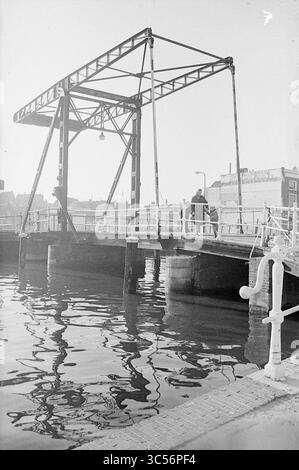 Report NZH Leiden, Noord Zuid Hollandse Vervoersmaatschappij N.V., NZ, 25-11-1970 Whizgle News, Dutch Desk, Paesi Bassi, 1950-2000 Un ponte oscillante si erge su acque calme, con la sua robusta struttura metallica che riflette i riflessi sottostanti. Una figura può essere vista camminare lungo il ponte, con edifici che fiancheggiano lo sfondo. La scena evoca un senso di tranquillità e fascino storico vicino al corso d'acqua. Foto Stock