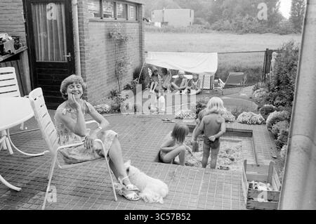 Donna con diversi bambini nel cortile, 00-07-1983 Whizgle News, Dutch Desk, Paesi Bassi, 1950-2000 Una donna siede su un patio, sorridendo alla macchina fotografica mentre si gode la giornata di sole. Sullo sfondo, un gruppo di bambini gioca vicino a un pozzo di sabbia e a una tenda improvvisata, circondata da vegetazione lussureggiante e arredi da giardino. Un piccolo cane siede accanto alla donna, aggiungendo all'atmosfera rilassata e allegra di una giornata estiva. Foto Stock