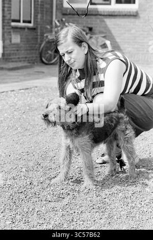Verticale. Donna con cane, 12-07-1967 Whizgle News, Dutch Desk, Paesi Bassi, 1950-2000 Una giovane donna in una top a righe accomoda giù, accarezzando delicatamente un cane arruffato. La scena si svolge all'aperto, con un accenno di una bicicletta visibile sullo sfondo, suggerendo un momento informale e sereno condiviso tra di loro. Foto Stock