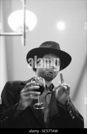 Uomo ritratto, 20-03-1991 Whizgle News, Dutch Desk, Paesi Bassi, 1950-2000 Un uomo con un elegante cappello regge un bicchiere di vino rosso, facendo una posa giocosa con un dito sollevato, suggerendo un momento spensierato. La sua espressione è un mix di curiosità e maltrattamento, che aggiunge fascino alla scena. Foto Stock