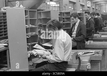 Smistamento della posta, PTT, servizi postali, posta cittadina, 22-01-1970 Whizgle News, Dutch Desk, Paesi Bassi, 1950-2000 Un gruppo di persone è impegnato nello smistamento attraverso pile di lettere e documenti in un ampio spazio di lavoro, con slot di posta organizzati in background. L'atmosfera suggerisce un ambiente vivace incentrato sull'efficienza e l'organizzazione. Foto Stock