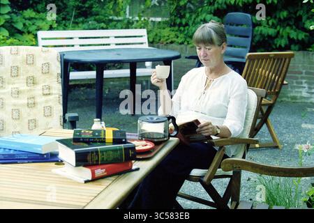 Mieke van Santen, Vijfhuizen, Vijfhuizen, 21-07-2003 Whizgle News, Dutch Desk, i Paesi Bassi, 1950-2000 Una donna siede comodamente a un tavolo da patio, gustando una tazza di caffè mentre legge un libro, circondato da una selezione di altri libri e da alcuni oggetti sparsi. La vegetazione lussureggiante offre uno sfondo sereno al suo momento di pace. Foto Stock