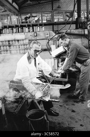 Visita soffiatura del vetro, vetrate colorate, vetrate soffiate, vetrate in generale, 14-06-1967 Whizgle News, Dutch Desk, Paesi Bassi, 1950-2000 due uomini sono impegnati in un processo di lavorazione del vetro all'interno di una fabbrica. Un uomo, vestito con un cappotto bianco e occhiali, maneggia con cura uno stampo metallico, mentre l'altro assiste nelle vicinanze, concentrandosi sul suo compito. L'atmosfera è laboriosa, con attrezzature visibili e uno spazio di lavoro pieno di prodotti in vetro sullo sfondo. Foto Stock