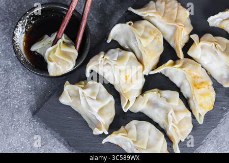 Un piatto di gyoza cotti, con uno immerso in una piccola ciotola di salsa di soia con bacchette, presentati su una superficie ruvida scura. Foto Stock