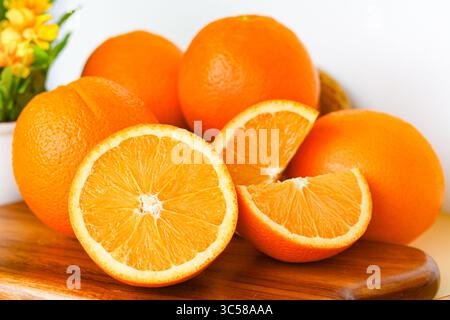 Primo piano di un gruppo di arance sunkiste intere e tagliate a fette disposte su un tagliere di legno e in un cesto, con un piccolo vaso di fiori sullo sfondo. Foto Stock