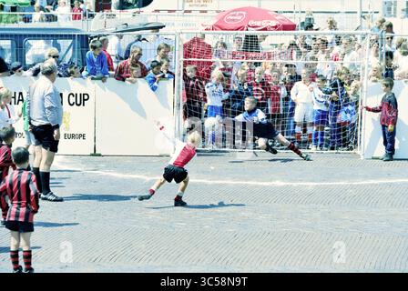 Torneo di calcio di strada, 25-07-1996 Whizgle News, Dutch Desk, Paesi Bassi, 1950-2000 Un giovane giocatore in rosso tenta un calcio di rigore mentre il portiere in immersioni nere per fare un salvataggio. La scena è piena di spettatori impazienti, mostrando un'atmosfera vivace in un evento di calcio giovanile. Foto Stock