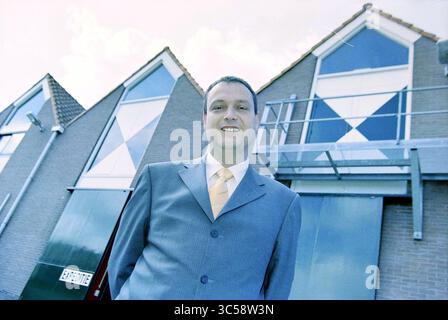Hans van de Boer, Cavex, Haarlem [Hans Maarten Den Boer], Haarlem, Paesi Bassi, 15-07-2001 Whizgle News, Dutch Desk, paesi Bassi, 1950-2000 Un uomo con un abito sartoriale si erge con sicurezza davanti a edifici moderni caratterizzati da sorprendenti design geometrici. Il suo sorriso amichevole completa l'architettura contemporanea dietro di lui. Foto Stock