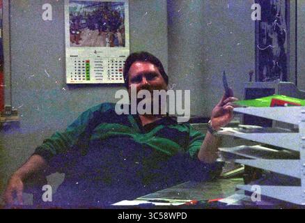 Ritratto di un uomo Whizgle News, Dutch Desk, Paesi Bassi, 1950-2000 Un uomo vestito in modo informale si siede rilassato alla sua scrivania, tenendo una penna e indossando un leggero sorriso. Dietro di lui, un calendario e varie forniture per ufficio suggeriscono un'area di lavoro piena di attività. Foto Stock