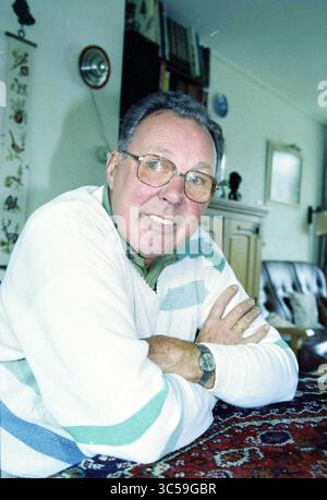 Ritratto di un uomo Whizgle News, Dutch Desk, Paesi Bassi, 1950-2000 un uomo anziano con gli occhiali e un sorriso amichevole siede comodamente con le braccia incrociate, circondato da un accogliente spazio abitativo che include scaffali e un muro decorativo. Foto Stock