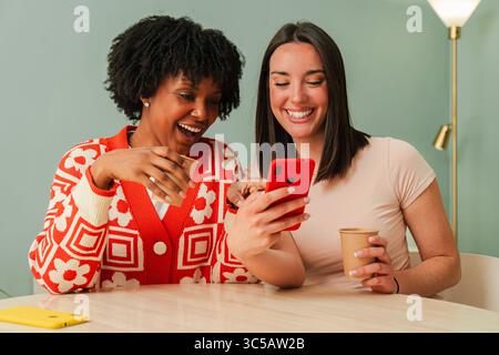 Due allegri femmine si sono riunite eccitatamente, affascinate da uno smartphone mentre guardano un video divertente, condividendo risate e gioia Foto Stock