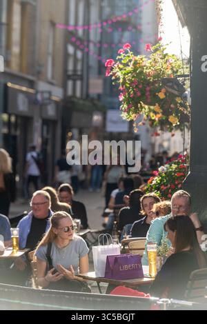 Persone che amano bere e fare shopping all'aperto in un pomeriggio di sole a Soho, Londra, con borse Liberty e vivaci esposizioni floreali Foto Stock