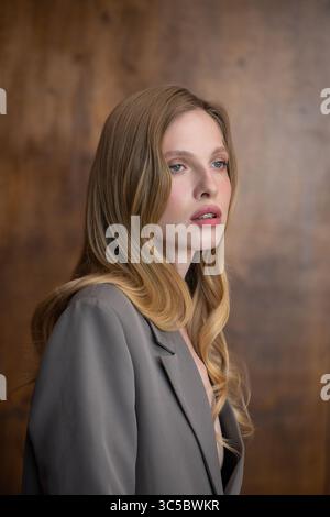 Una bella giovane donna dai lunghi capelli ondulati biondi, che indossa un elegante blazer grigio, guarda con attenzione lateralmente su uno sfondo caldo e sfocato Foto Stock