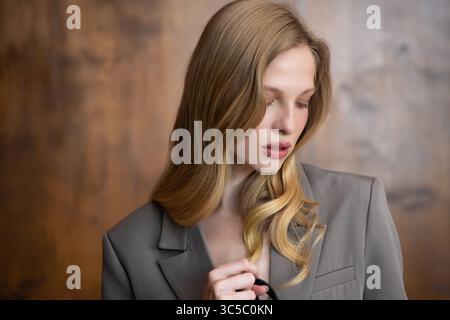 Una bella giovane donna dai lunghi capelli ondulati biondi, che indossa un elegante blazer grigio, guarda con attenzione lateralmente su uno sfondo caldo e sfocato Foto Stock