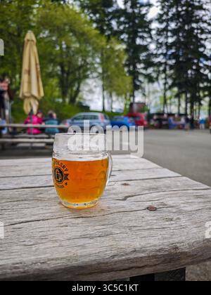 Bicchiere di birra su un rustico tavolo di legno all'aperto, con sfondo sfocato di auto, persone e alberi in un pub o birreria di campagna. Foto Stock