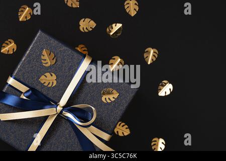 Frammento di scatola regalo blu con nastro in raso oro e blu. Sfondo nero, coriandoli dorati a forma di foglia di monstera, spazio per il testo. Foto Stock