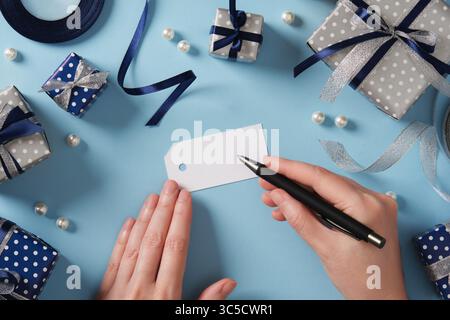 Le mani firmano un cartellino regalo. Circondato da regali e nastri nei colori blu e argento. Prepararsi per le vacanze. Spazio per il testo sull'etichetta. Foto Stock