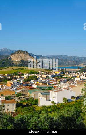 I tetti di Cuevas de San Marcos conducono lo sguardo verso la ripida collina coronata da alberi e la diga del bacino idrico di Iznajar, con montagne e ulivi gro Foto Stock