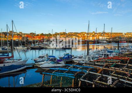 Vista mattutina con luce dorata sul porticciolo di Whitby, con barche ormeggiate e vasi di aragosta in primo piano e in collina sullo sfondo Foto Stock