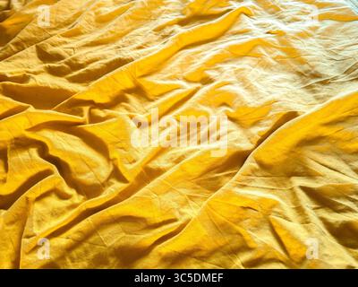 La luce soffusa e calda del sole illumina un lenzuolo giallo dorato ondulato, creando uno sfondo dalle texture invitanti e astratte con pieghe e rughe delicate Foto Stock