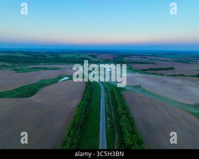 Una veduta aerea cattura una lunga strada rettilinea che attraversa ampi campi nebbiosi, che conduce verso villaggi e foreste lontani sotto una mornina morbida Foto Stock