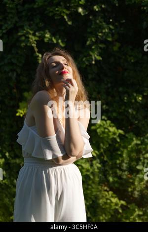Una giovane donna si erge graziosamente all'aperto con un top e pantaloni bianchi a spalla, circondati da una vegetazione lussureggiante Foto Stock