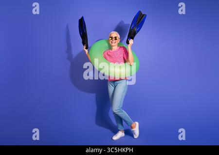 Donna anziana gioiosa in un abbigliamento elegante con pinne verdi gonfiabili e pinne da snorkeling su sfondo viola che simboleggiano la vibrante energia estiva Foto Stock