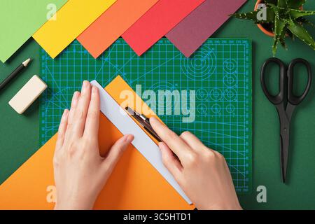 Taglio manuale di carta colorata con un taglierino sul tappetino da taglio verde nella vista dall'alto. Processo creativo, preparazione di progetti realizzati a mano. Foto Stock