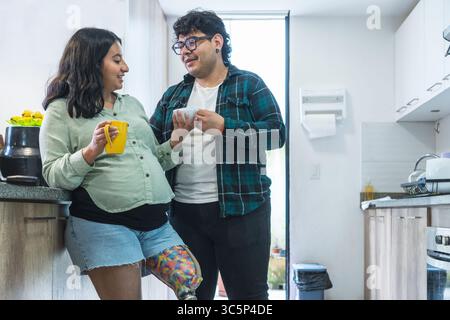 Coppia felice, mamma incinta con gamba protesica e marito, che condividono un momento insieme mentre bevono un caffè nella loro cucina Foto Stock