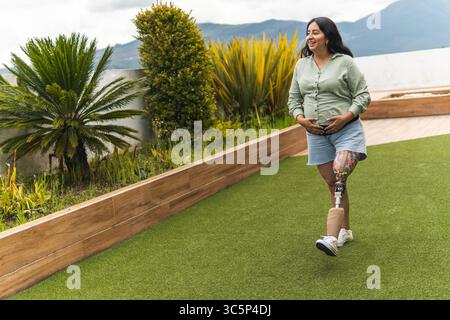 Donna incinta felice con una gamba protesica che cammina e tocca la pancia in un giardino Foto Stock