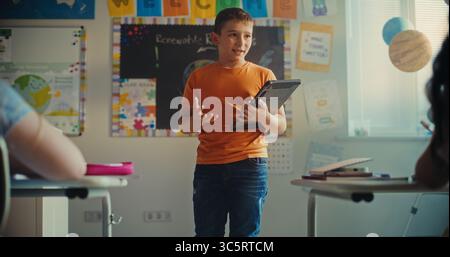 Classe STEM sull'energia rinnovabile: Scuola elementare ragazzo con tablet computer che presenta i compiti sull'ecologia davanti ai compagni di classe. Diversi bambini seduti alle scrivanie durante la lezione di scienze in classe. Foto Stock
