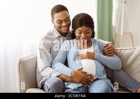 Uomo sorridente che abbraccia la sua donna incinta, seduto sul divano Foto Stock