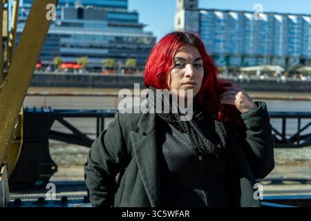 Giovane donna con capelli rossi e abiti moderni in posa a puerto madero, con le banchine e gli edifici sullo sfondo Foto Stock