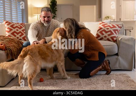 Coppia che indossa maglioni in maglia inclinati in avanti accarezzando Golden retriever sul divano grigio del soggiorno Foto Stock