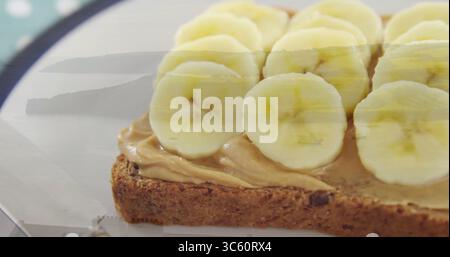 Visualizzazione del pane tostato a grani interi con burro di arachidi e fette di banana sul piatto decorativo con coltello Foto Stock