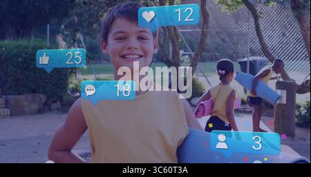 Ragazzo sorridente in cima gialla che porta con sé un tappetino da yoga blu in un parco soleggiato, con icone di reazione galleggianti Foto Stock