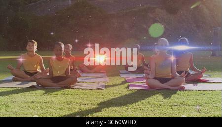 Meditare i bambini seduti a gambe incrociate su tappetini da yoga nel parco su un prato erboso, con le cime gialle Foto Stock