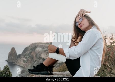 Donna laptop Cliffside Sunset - Una donna siede su una scogliera con un notebook, guardando il tramonto. Foto Stock