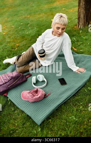 In un parco lussureggiante, una giovane donna alla moda si rilassa su una coperta, gustando il suo caffè e rilassandosi. Foto Stock
