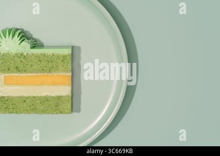 Fetta di torta bento colorata su un piatto verde chiaro, rivelando strati di pistacchio verde, panna, mango e frutto della passione, sottolineando l'incrocio artistico Foto Stock