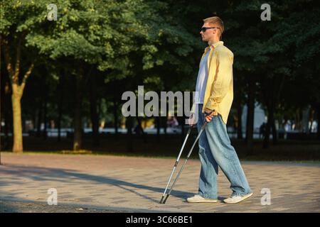 Uomo con disabilità visiva che cammina su un sentiero lastricato con bastone nel parco cittadino Foto Stock
