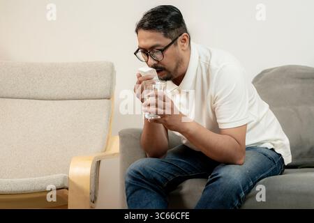 Un giovane malato si siede a casa prendendo la medicina della febbre, rappresentando l'autoassistenza e gestendo i sintomi comuni della malattia. Foto Stock