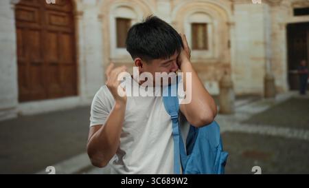 Un giovane uomo in una strada cittadina con uno zaino blu che sembra stressato, all'aperto suggerisce uno studente che affronta sfide o emozioni, sottolineando un urb Foto Stock