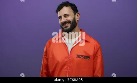 Uomo ispanico in una tuta di prigione arancione sorridente con fiducia su un vibrante sfondo viola. Foto Stock