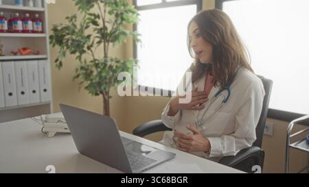 Medico ispanico femminile in stanza d'ospedale che conduce videochiamata utilizzando il computer portatile, con stetoscopio intorno al collo, trasmettendo professionalità in clinica in interni Foto Stock