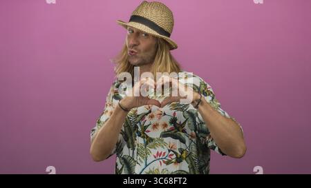 Uomo che forma il cuore con le mani sorridente e indossa cappello di paglia e camicia tropicale in studio rosa; affetto. Foto Stock