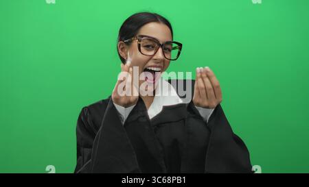 Giudice donna sorridente in veste nera facendo gesto di soldi su un muro verde isolato che esprime successo finanziario o ambizione Foto Stock