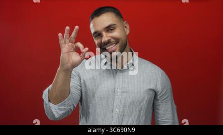 Uomo che fa a mano il segno con entrambe le mani e sorridente mentre indossa una camicia casual in un luminoso ambiente da studio rosso; approvazione gioia. Foto Stock