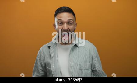 Giovane uomo che esprime gioia su un vivace sfondo arancione indossando una camicia grigia casual e sorridendo ampiamente e coinvolgendo gli spettatori con energia e unicità Foto Stock