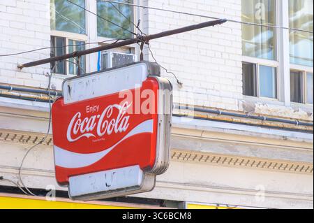 Un'insegna vintage rossa e bianca della Coca-Cola è appesa a un'asta intemprata su un edificio in mattoni bianchi con vecchie finestre sopra. Foto Stock